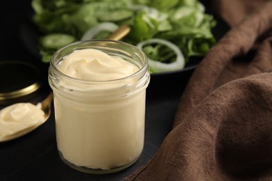 Photo of Jar and spoon with delicious mayonnaise near fresh salad on black table