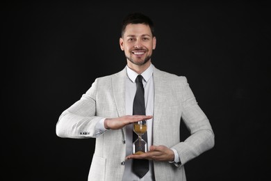 Happy businessman holding hourglass on black background. Time management