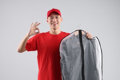 Photo of Dry-cleaning delivery. Happy courier holding garment cover with clothes and showing OK gesture on light grey background