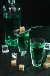 Photo of Absinthe in shot glasses, spoon, brown sugar and ice cubes on gray table against dark background, closeup. Alcoholic drink