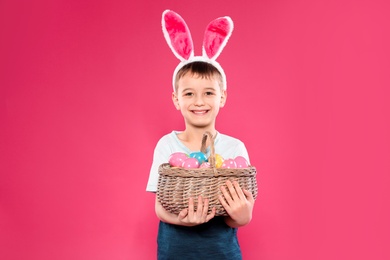 Photo of Little boy in bunny ears headband holding basket with Easter eggs on color background