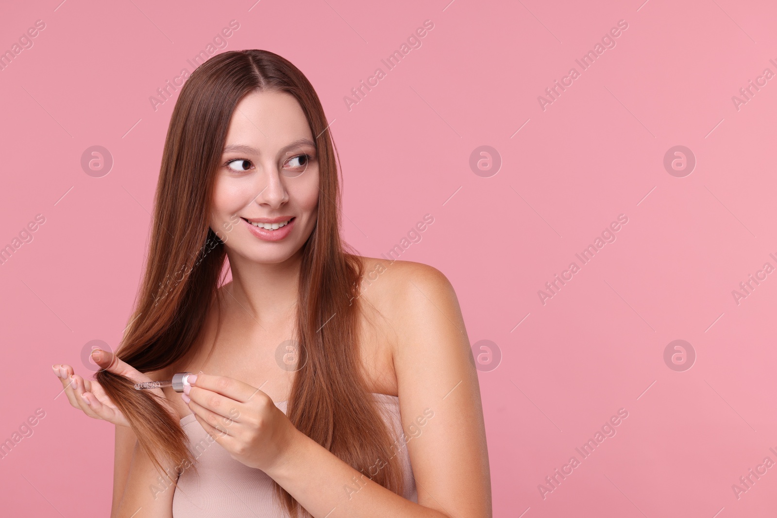 Photo of Beautiful woman applying serum onto hair on pink background, space for text