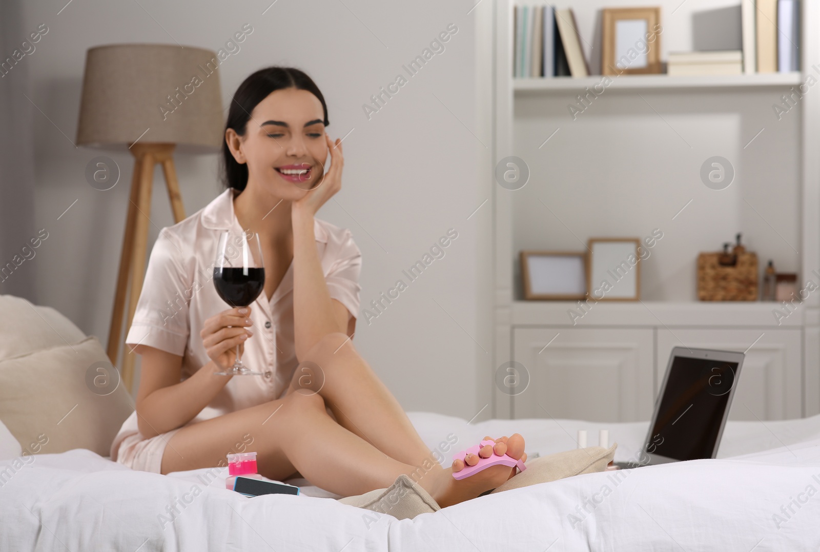 Photo of Beautiful young woman with glass of wine giving herself pedicure on bed at home