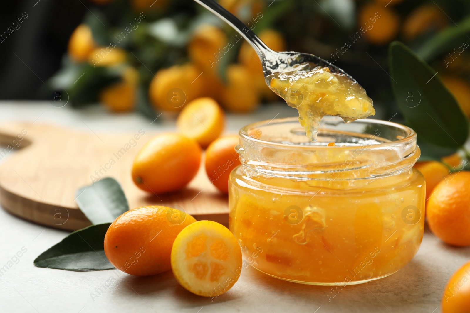 Photo of Spoon with delicious kumquat jam above jar and fresh fruits on light grey table, space for text