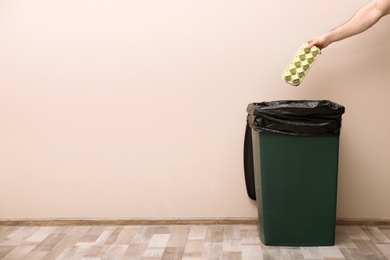 Photo of Young woman throwing egg carton in trash bin indoors, space for text. Waste recycling