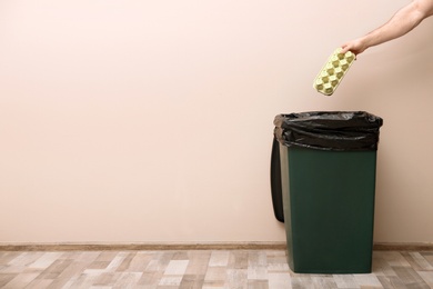 Young woman throwing egg carton in trash bin indoors, space for text. Waste recycling