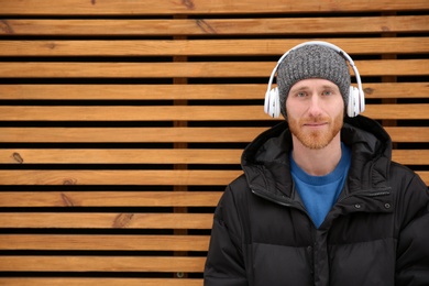 Photo of Young man listening to music with headphones against wooden wall. Space for text
