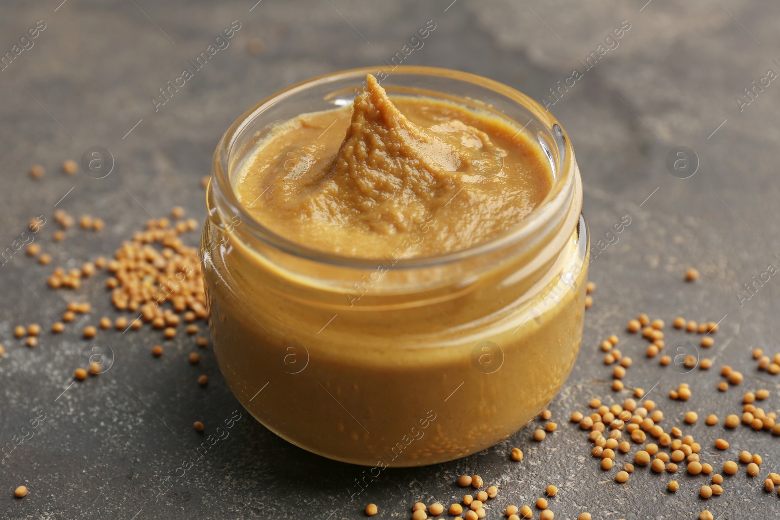 Photo of Tasty mustard sauce in jar and dry seeds on grey textured table, closeup