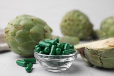 Bowl with pills and fresh artichokes on white tiled table, closeup