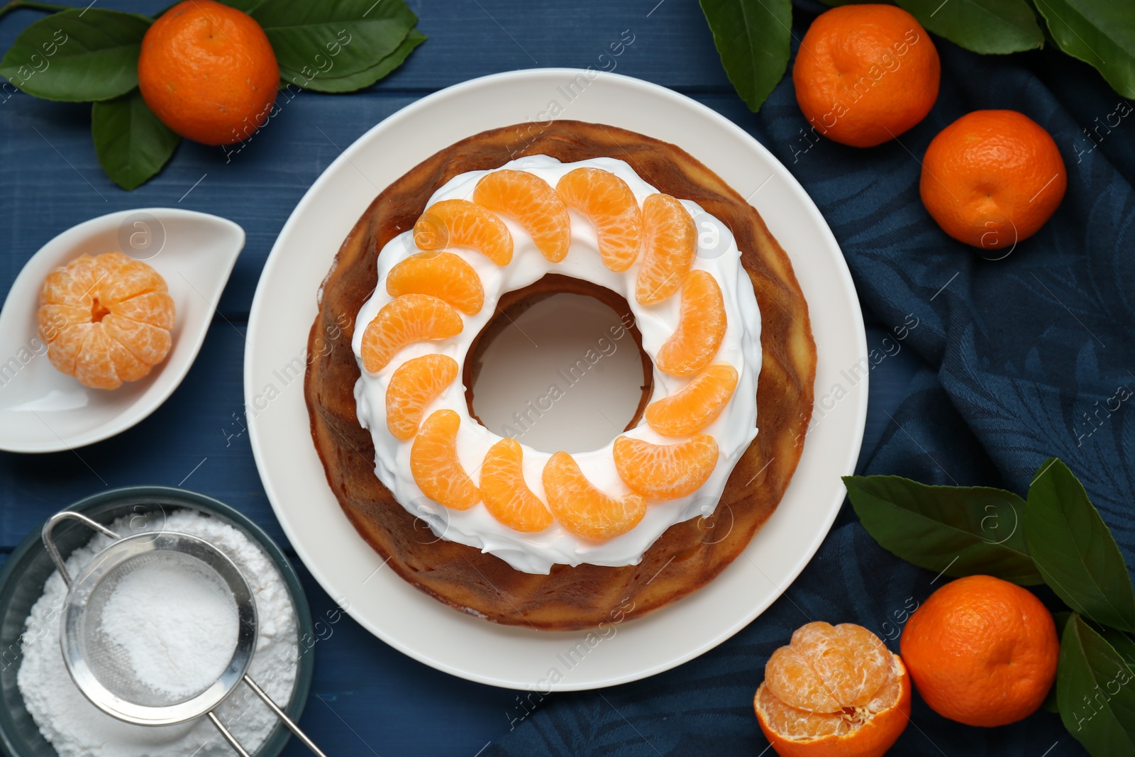 Photo of Homemade yogurt cake with tangerines, cream and green leaves on blue wooden table, flat lay