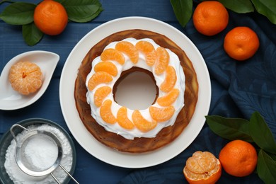 Homemade yogurt cake with tangerines, cream and green leaves on blue wooden table, flat lay