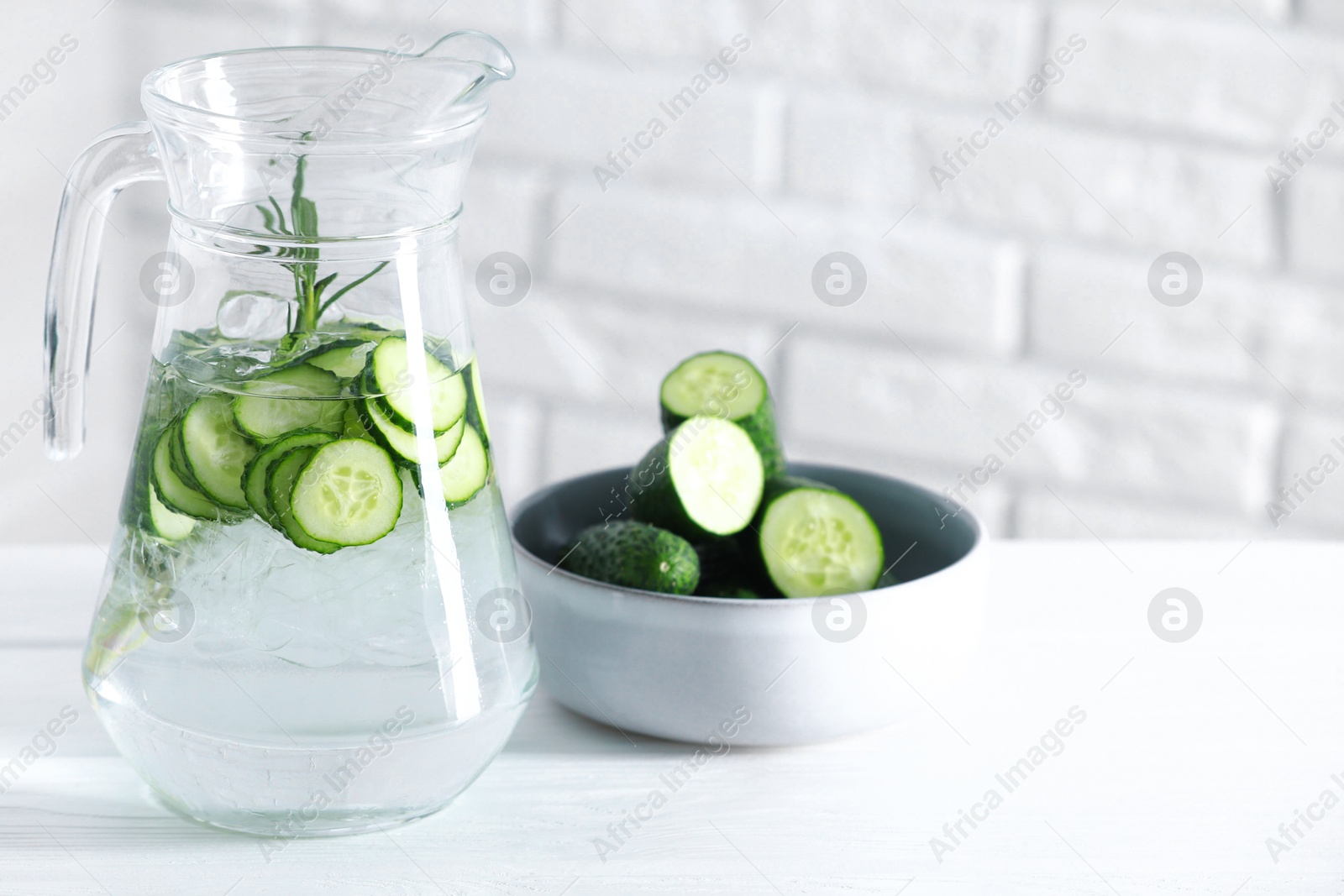 Photo of Refreshing cucumber water with rosemary in jug and vegetables on white table against brick wall, closeup. Space for text