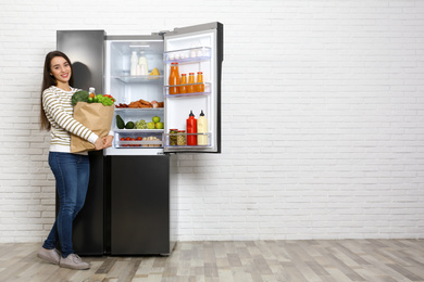 Young woman with paper bag full of products near open refrigerator indoors, space for text