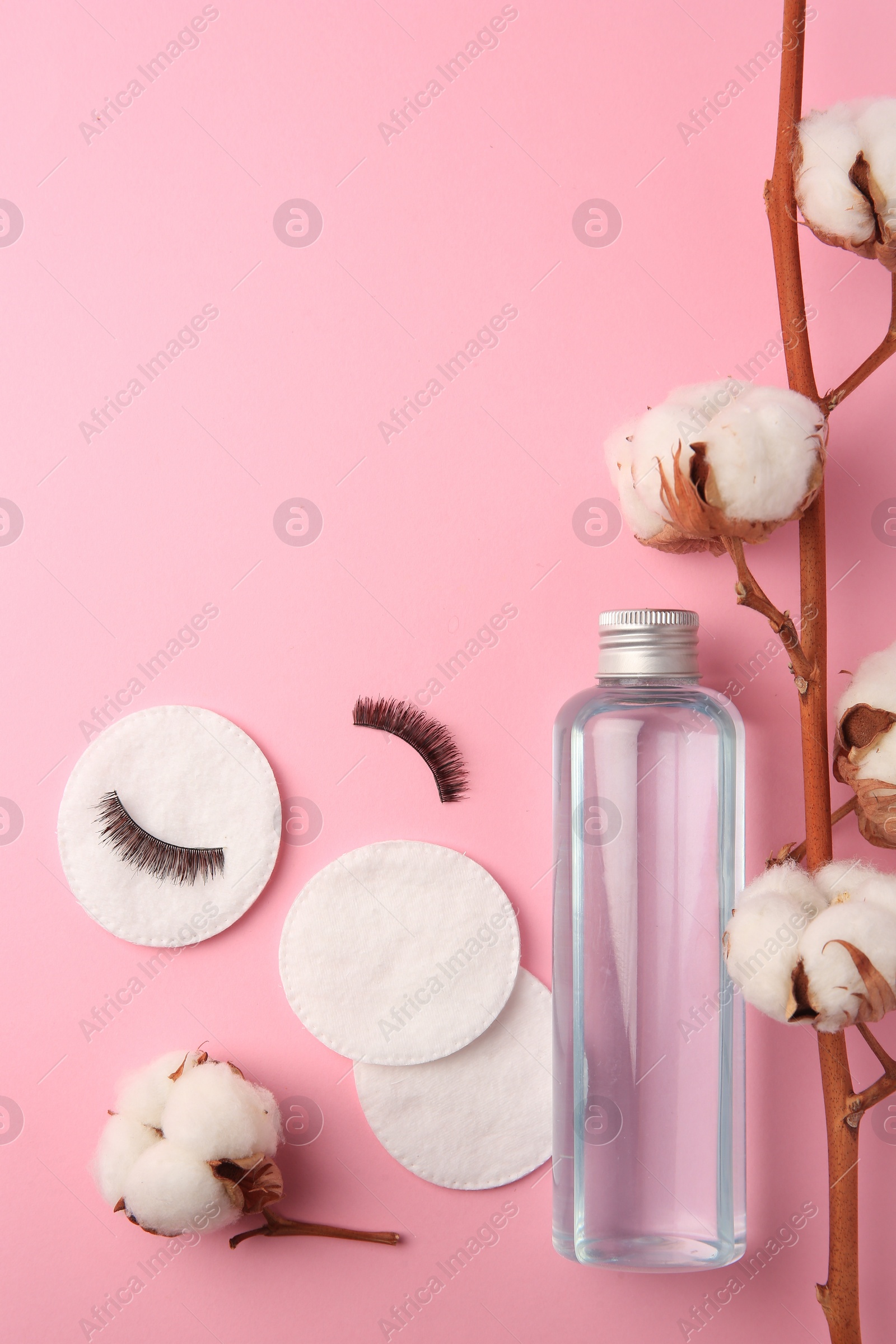 Photo of Bottle of makeup remover, cotton flowers, pads and false eyelashes on pink background, flat lay. Space for text