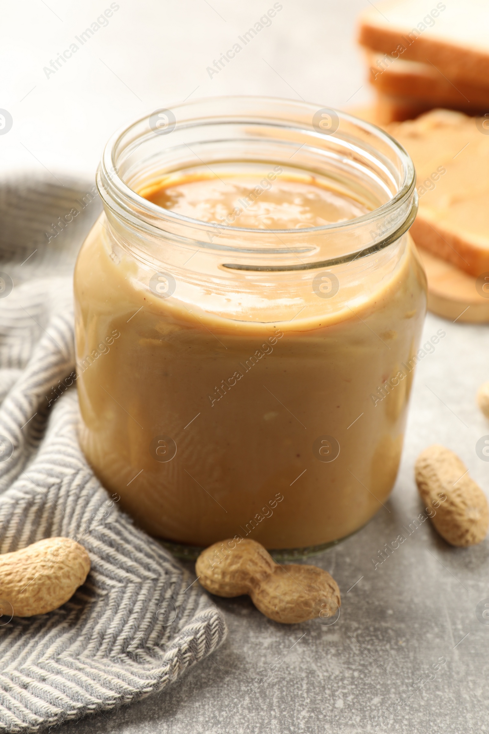 Photo of Tasty peanut butter in glass jar and peanuts on gray table, closeup