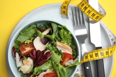 Photo of Plate of tasty salad, cutlery and measuring tape on yellow background, top view. Nutrition regime