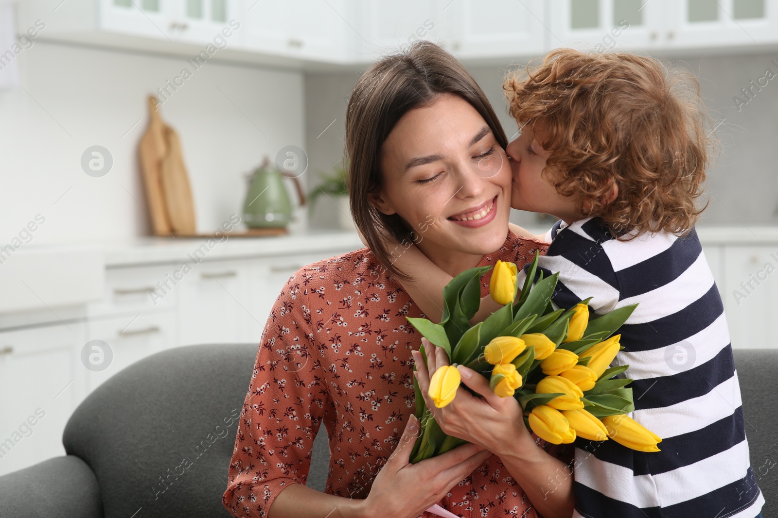 Photo of Little son kissing and congratulating his mom with Mother`s day at home. Woman holding bouquet of yellow tulips