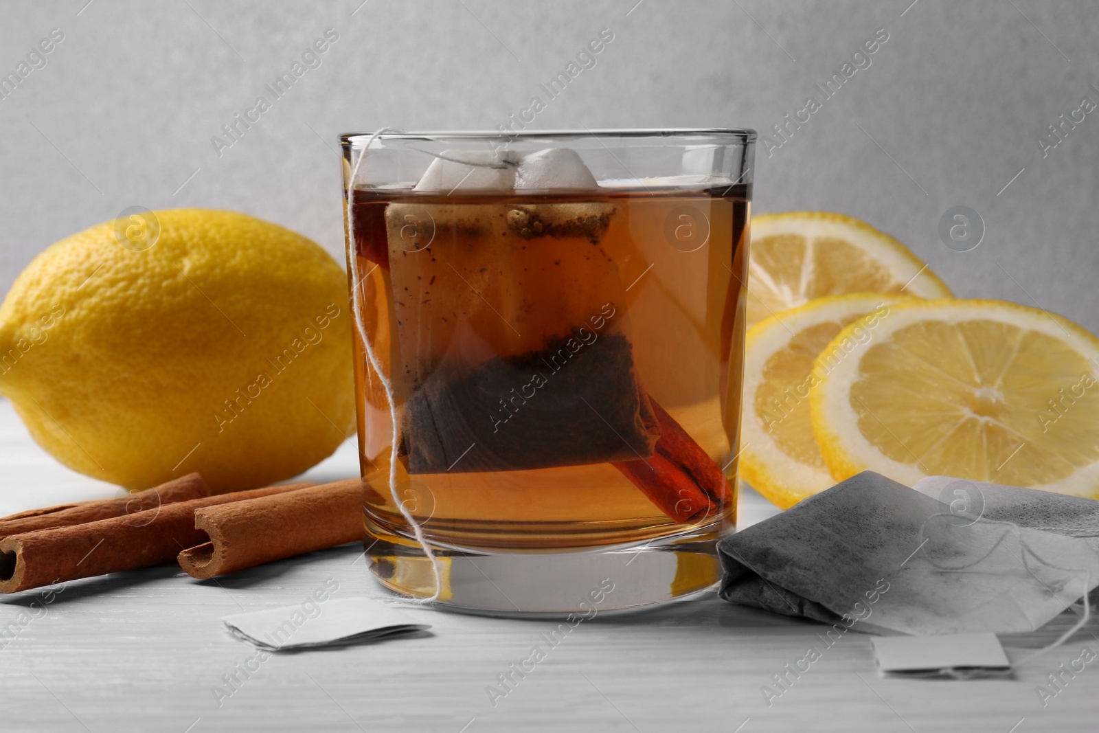 Photo of Tea bags, glass of hot drink, cinnamon sticks and lemons on white wooden table, closeup