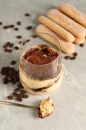 Photo of Tasty tiramisu in glass, coffee beans, spoon and biscuits on light grey table, closeup