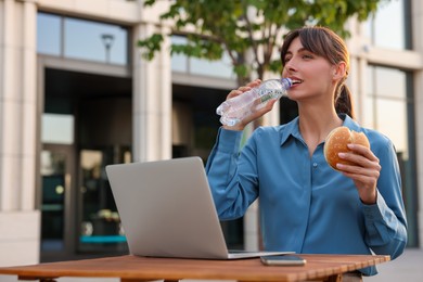 Businesswoman with hamburger drinking water from bottle during lunch at wooden table outdoors