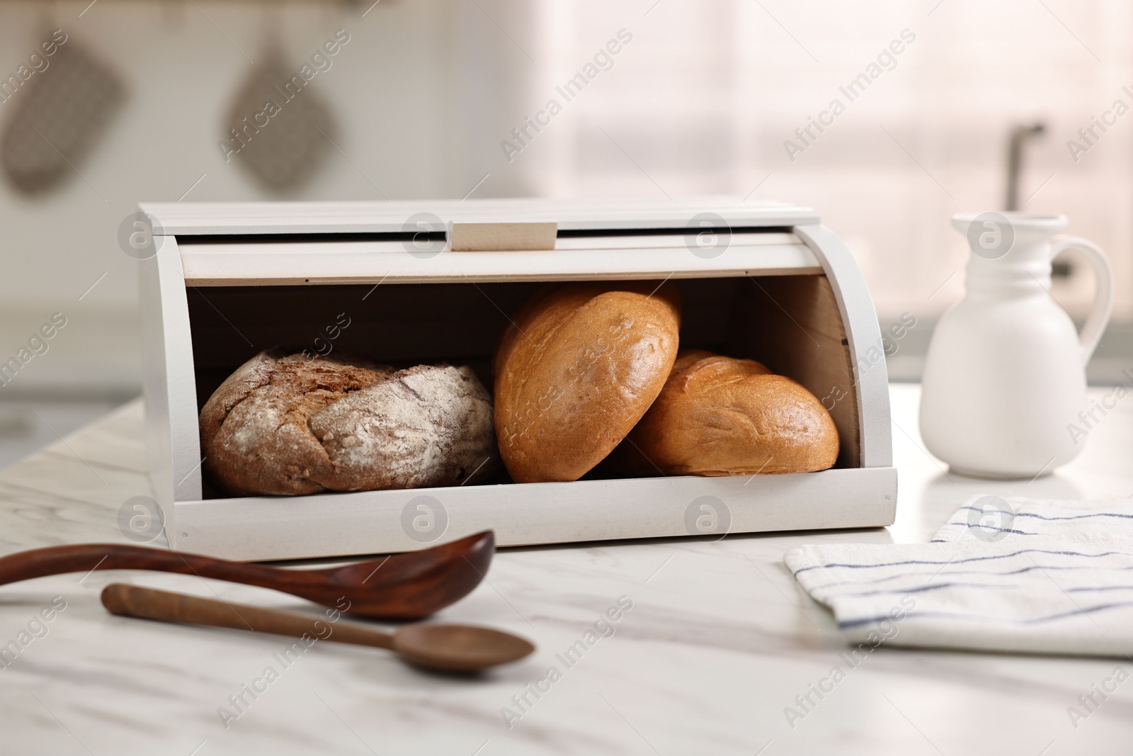Photo of Wooden bread basket with freshly baked loaves and spoons on white marble table in kitchen
