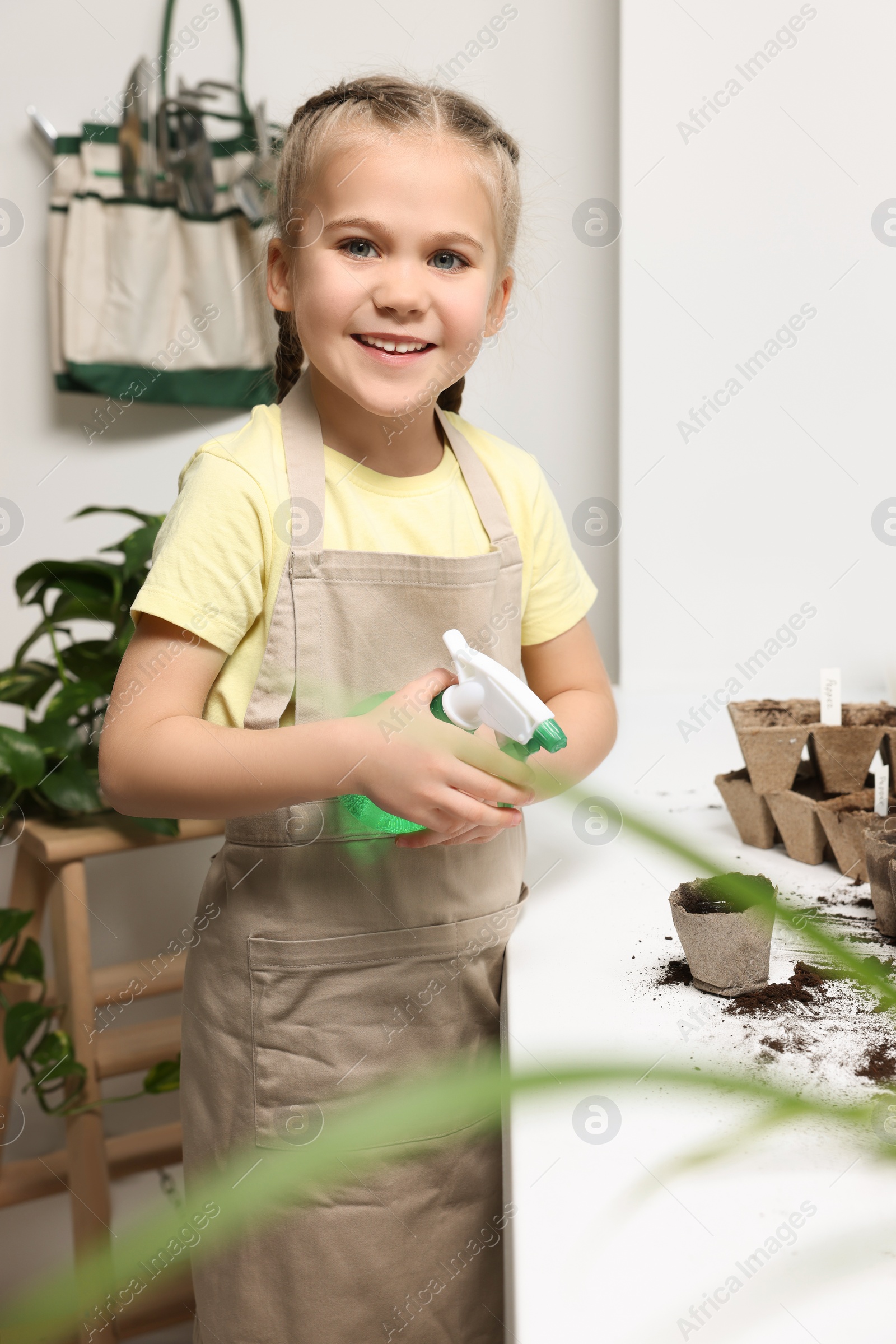 Photo of Little girl spraying water onto vegetable seeds in peat pots on window sill indoors