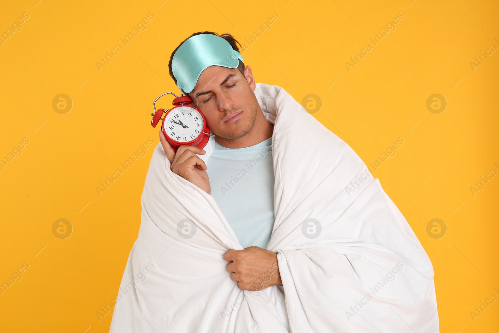 Photo of Man in sleeping mask wrapped with blanket holding alarm clock on yellow background