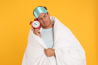 Photo of Man in sleeping mask wrapped with blanket holding alarm clock on yellow background