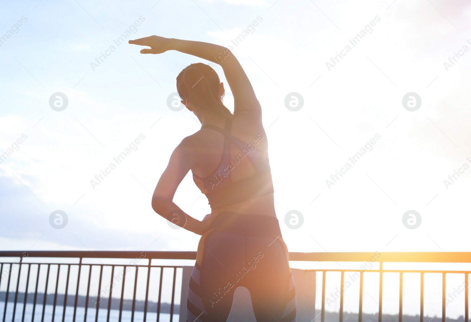 Photo of Young woman stretching on pier near river in morning, back view