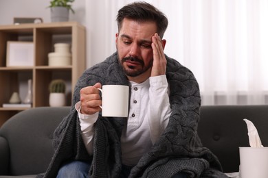 Sick man wrapped in blanket with cup of drink on sofa at home. Cold symptoms