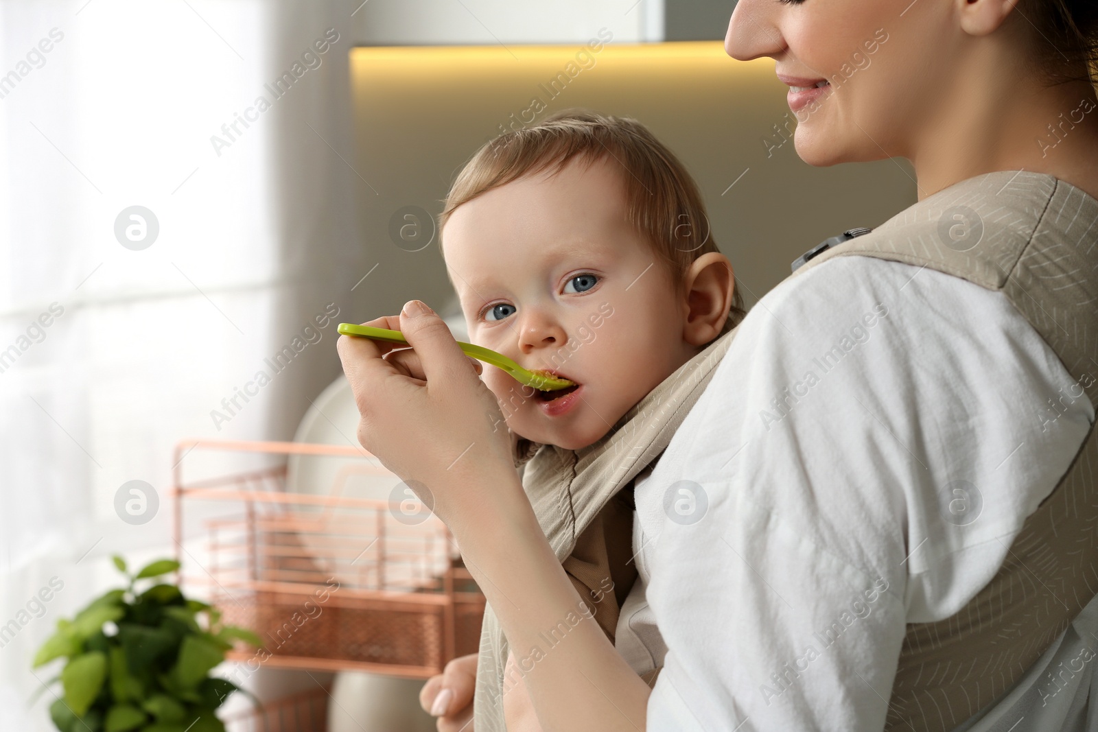 Photo of Mother feeding her cute child in sling (baby carrier) indoors