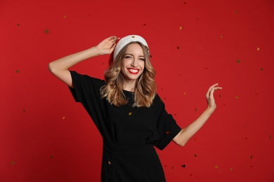 Photo of Happy young woman wearing Santa hat and confetti on red background. Christmas celebration