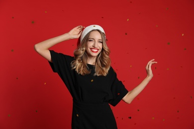 Happy young woman wearing Santa hat and confetti on red background. Christmas celebration