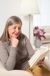 Beautiful senior woman reading book in armchair at home