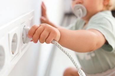 Photo of Cute baby playing with plug and electrical socket at home, closeup. Dangerous situation