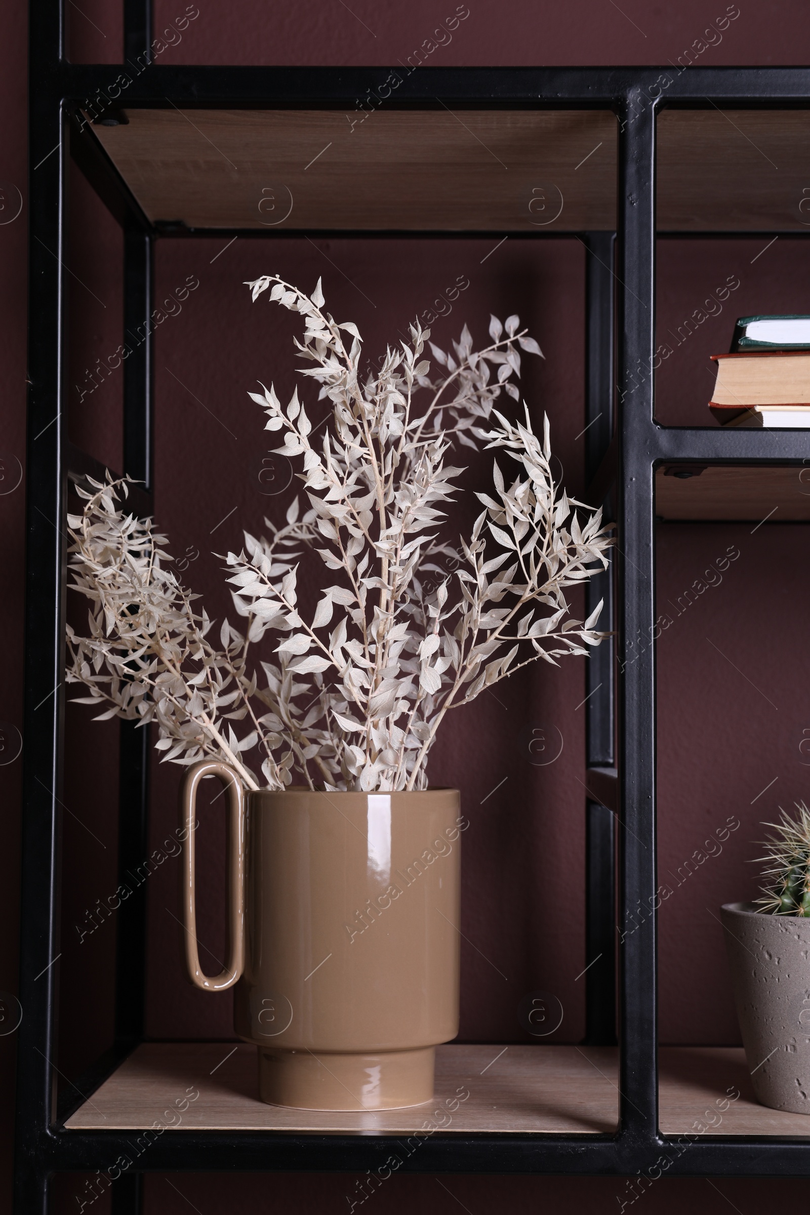 Photo of Stylish ceramic vase with dry plants on shelf near brown wall