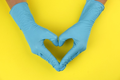 Person in medical gloves making heart with hands on yellow background, top view