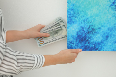 Photo of Woman hiding dollar banknotes behind painting indoors, closeup. Money savings