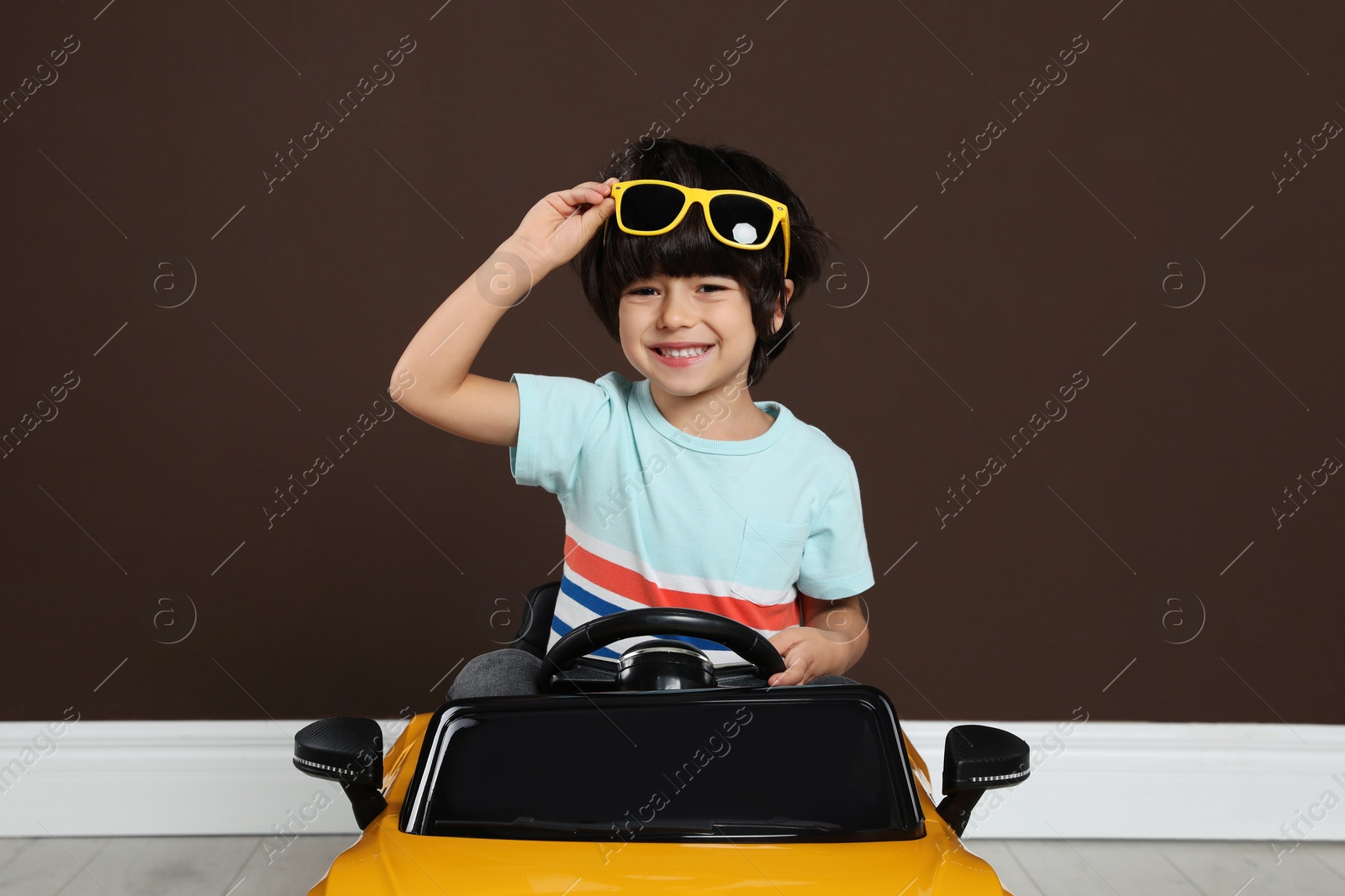 Photo of Cute little boy driving children's electric toy car near brown wall indoors