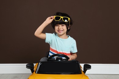 Cute little boy driving children's electric toy car near brown wall indoors