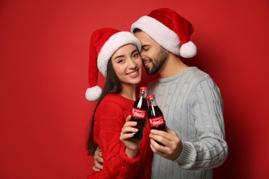 Photo of MYKOLAIV, UKRAINE - JANUARY 27, 2021: Young couple in Christmas hats holding bottles of Coca-Cola on red background