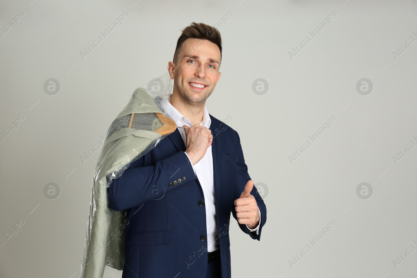 Photo of Man holding hanger with jacket in plastic bag on light grey background, space for text. Dry-cleaning service