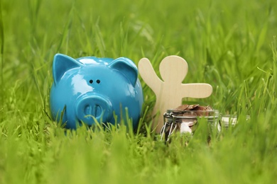 Photo of Cute piggy bank, wooden person and jar with coins on green grass in park