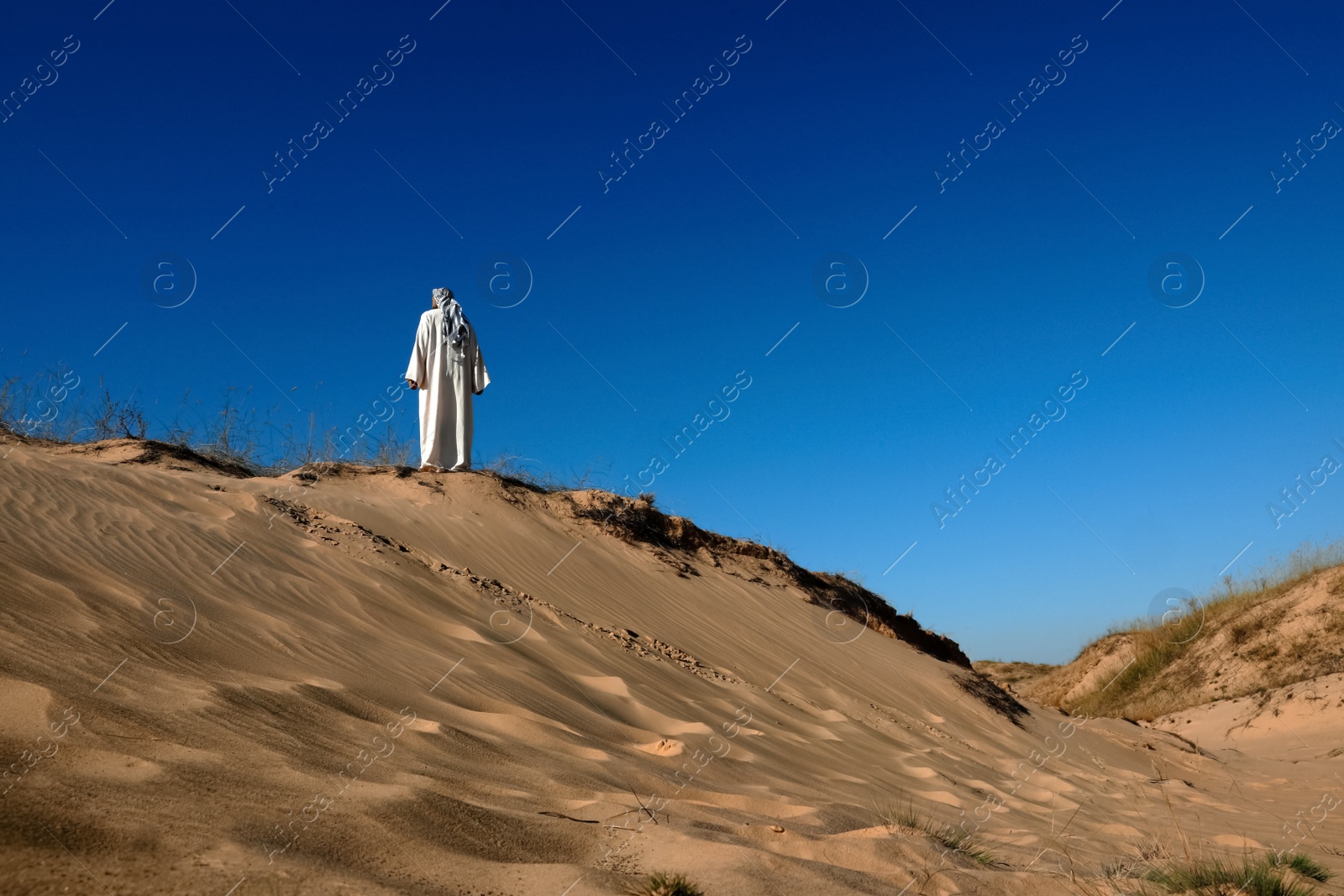 Photo of Man in arabic clothes walking through desert on sunny day, back view