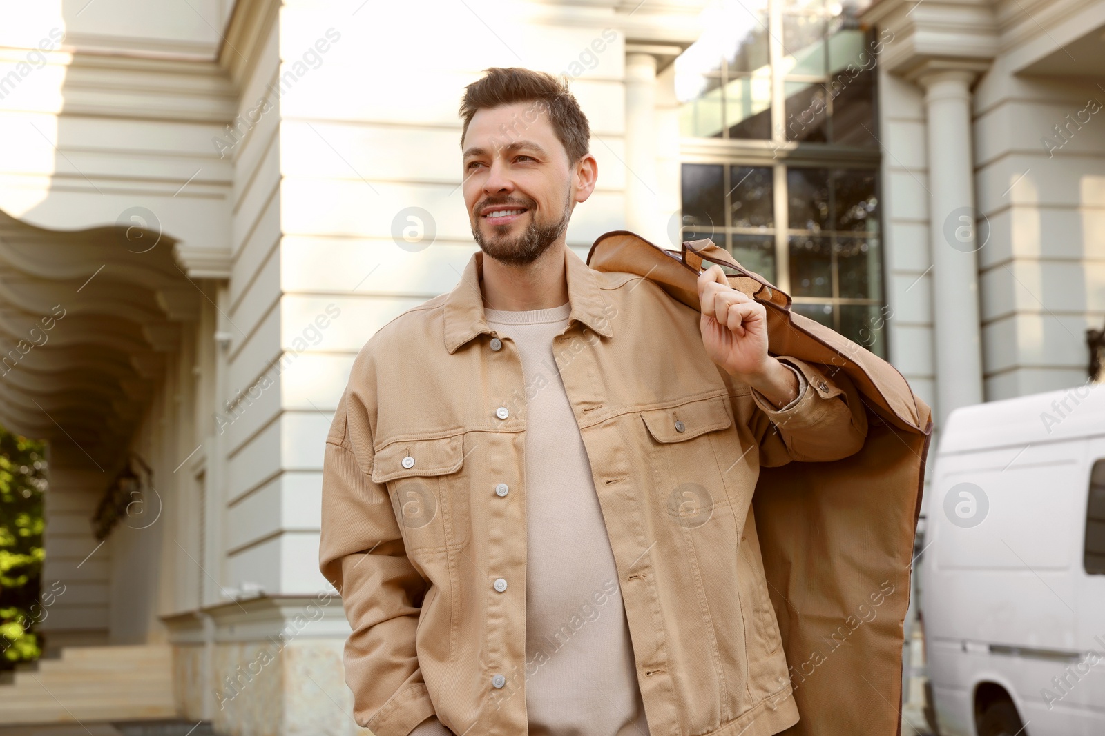 Photo of Attractive happy man holding garment cover with clothes outdoors. Dry cleaning service