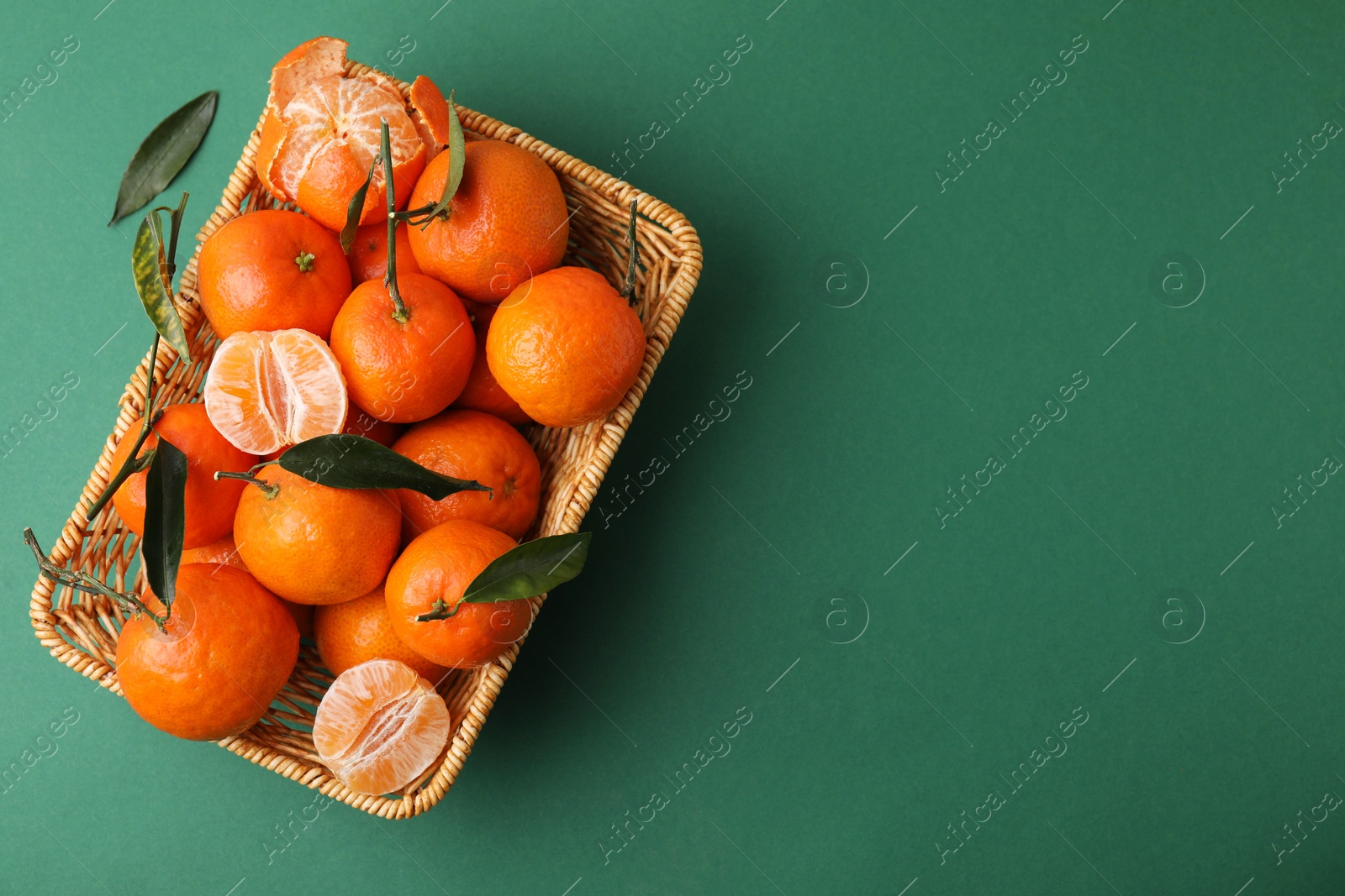 Photo of Fresh ripe tangerines with leaves in wicker basket on green background, top view. Space for text