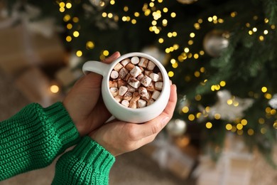 Photo of Woman holding cup of delicious Christmas cocoa with marshmallows near fir tree, top view