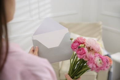 Photo of Woman holding envelope with blank greeting card and bouquet of ranunculus at home, closeup