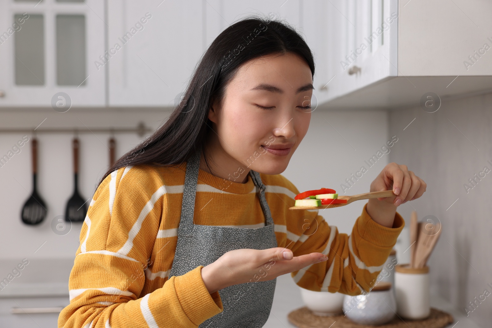 Photo of Beautiful woman cooking and tasting vegetable dish in kitchen