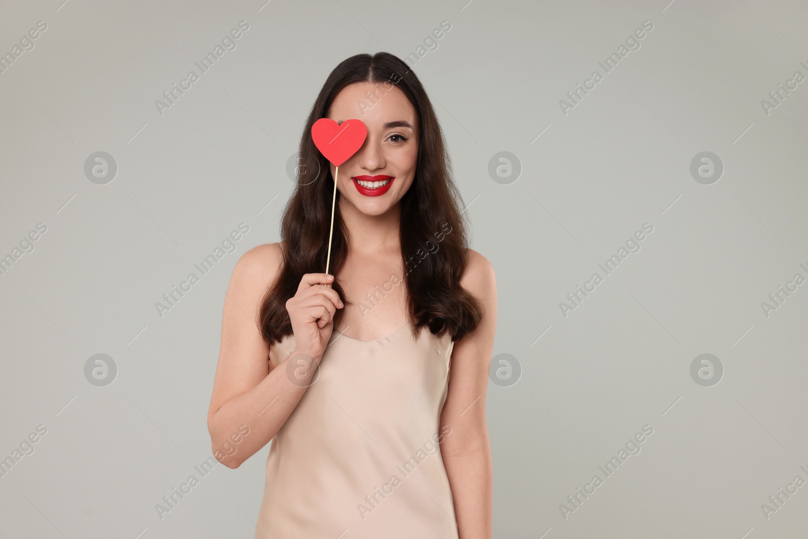 Photo of Beautiful young woman covering her eye with paper heart on grey background